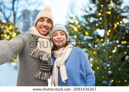 people and winter holidays concept - happy couple in knitted hats and scarves taking selfie outdoors over christmas tree lights background
