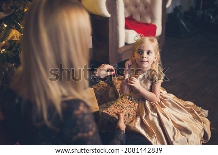 A little girl in an elegant dress and her mom open a gift box under the Christmas tree.