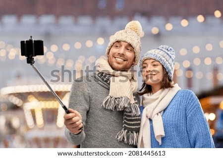 people, technology and winter holidays concept - happy couple in knitted hats and scarves taking picture by smartphone on selfie stick over christmas market and lights background