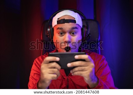 Playing video game on smartphone. Young asian handsome man sitting on chair holding cellphone in living room. Happiness Streamer Indian man wearing headset playing game online in the darkroom.