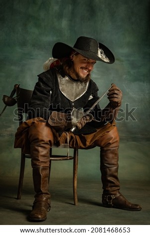 Brutal horrendous man, pirate in vintage medieval costume sitting and holding sword isolated over dark background. Combination of medeival and modern styles. Concept of history. Copyspace for ad.