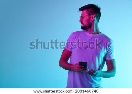 News. Portrait of man holding phone and inquiringly looking leftside isolated over blue background in neon lights. Concept of human emotions, facial expression, lifestyle. Copy space for ad