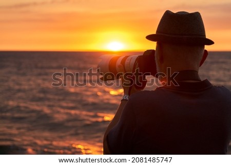  An adult man takes photos of the sunset over the sea with a camera. The sun sets below the horizon, the last rays of the sun are reflected in the water. Copy space.                              