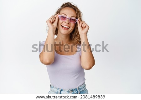 Happy young woman tries new sunglasses, smiles and looks pleased at camera, stands in stylish summer outfit against white background