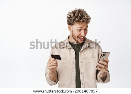 Excited blond man looking at his smartphone amazed, holding credit card, making money, staring at mobile application, standing over white background