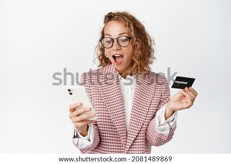 Amazed corporate woman in suit looking at her mobile phone, holding credit card, paying with smartphone, order online, standing against white background