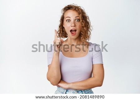 Surprised blond girl with curly hair making announcement, looking amazed, pointing finger up at advertisement, standing in tshirt over white background
