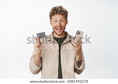Excited young man winning money on app, holding credit card and mobile phone, shopping online, standing in casual clothes over white background