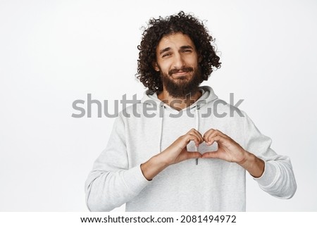 Image of heartfelt smiling young man, showing heart sign near chest, I love you gesture, looking caring at camera, white background
