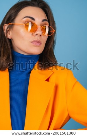 Fashionable young woman in a knee-length and oversized jacket on a blue background in stylish bright orange glasses posing