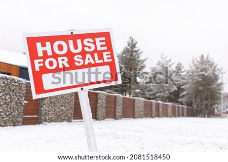 Red sign - house for sale, outdoors in winter against the backdrop of a fence and snowy pine trees. Buying suburban real estate with a mortgage. Sale of confiscated property. Moving to a new home.