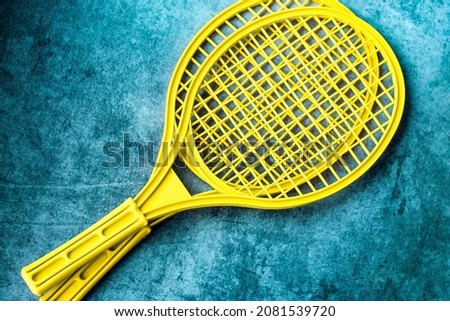 two plastic tennis rackets of bright yellow color are laid out diagonally on a worn turquoise background; the children's version of the tournament is badminton