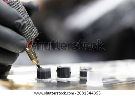 Detail of the tattooist's hand with black glove holding the tattoo machine while he loads ink into the needle while performing a tattoo. Image in horizontal and colour. Royalty-Free Stock Photo #2081544355
