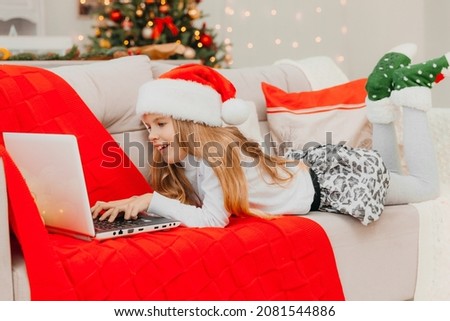 A little girl in a Santa Claus hat communicates by video call via a laptop, lying on the couch in the living room.