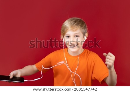 beautiful smiling boy on a red background holds a phone in his hand and listens to music through headphones