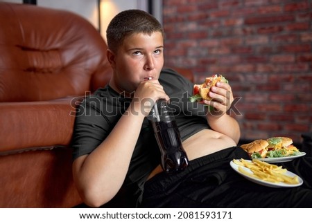 Overweight boy with fast food on floor near sofa at home, eating junk food drinking sweet beverages, lazy fat teenage boy in casual wear at home alone, having no control in eating behavior.