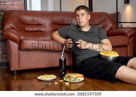 Boy is overeating. Fat child, food addiction and behavior problems. Lazy teenage boy on floor eating fast food alone after school, lead unhealthy lifestyle. overeating, obesity, overweight Royalty-Free Stock Photo #2081597509