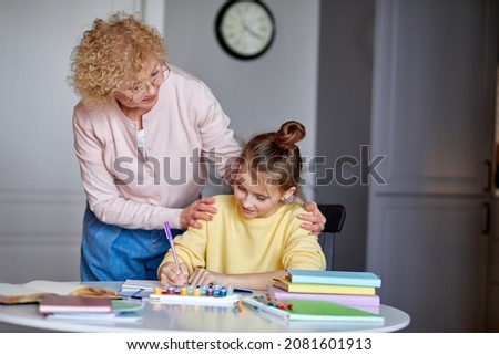 Creative activity. Happy mature grandma and preteen daughter friends sit at desk paint pictures in album using colored pencils. Focused little girl learn to draw with elderly female babysitter