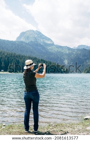 Woman stands on the shore and takes pictures of the mountains on her phone. Back view