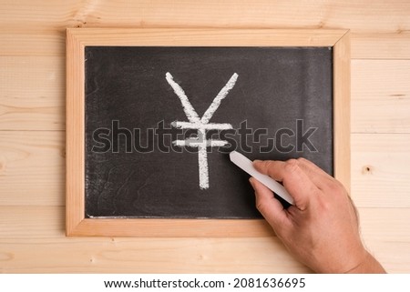 A man's hand draws a yen symbol on a chalkboard with white chalk.