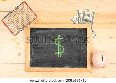 The Dollar symbol. A drawing of the dollar sign, on a black chalk board in a frame, next to dollar bills, a shopping basket and a piggy bank. Top view