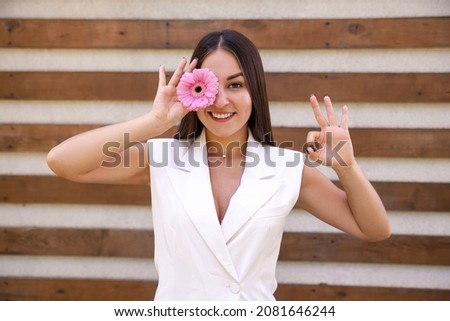 blossoms, emotions, gesture and people concept - portrait of an oriental girl in white clothing with a flower by the eye showing an okie sign with hands on a brown ribbed background with copy space