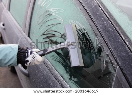 Frosted car side door window cleaning from a hoar frost before a drive at frozen winter day - Woman's hand with warm glove removes a white frost with silicone scraper with handle Royalty-Free Stock Photo #2081646619