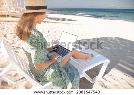 Rear view of an attractive young Caucasian woman in a green dress and hat sits on a lounger with a laptop in her hands in the shade of a wooden umbrella. Cut out laptop screen