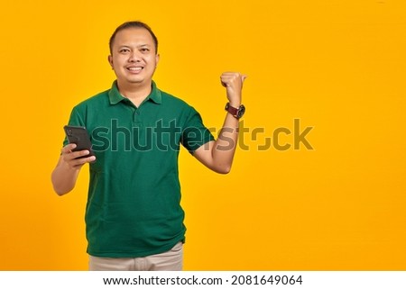 Cheerful young Asian man looking at mobile smartphone screen with open mouth on yellow background