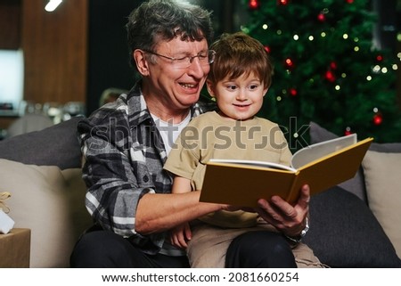 Mesmerized little boy and his grandpa sitting on a sofa looking through a photo album. Christmas tree in background.
