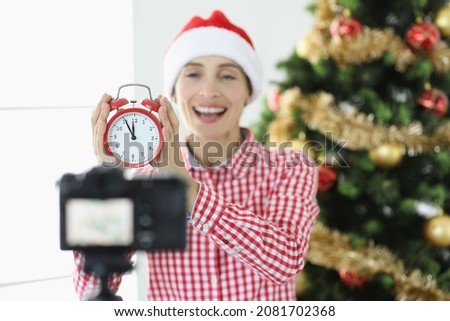 Joyful woman blogger shoots video on camera holding alarm clock on background of New Year tree concept