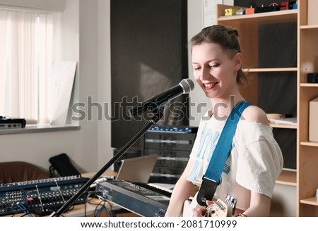 young woman musician playing guitar in studio during rehearsal. rock musician smiling and standing next to a microphone.