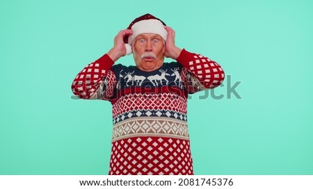 Oh my God Wow. Grandfather wears New Year sweater raising hands in surprise looking at camera with big eyes shocked by sudden victory isolated on light blue background studio. Happy Christmas holiday