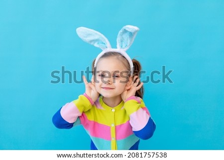 portrait of a little girl with bunny ears in a striped jacket smiling and indulging. The concept of the Easter holiday. Blue background, photo studio, place for text