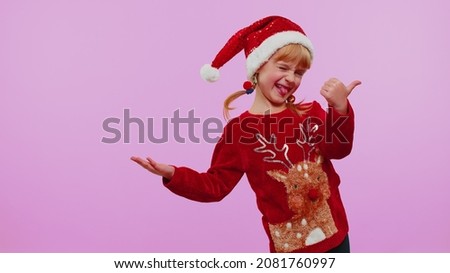 Portrait of funny kid girl in red Christmas sweater showing thumbs up and pointing at left on blank space. Place for your advertisement logo on violet background studio. Happy New Year holidays sale