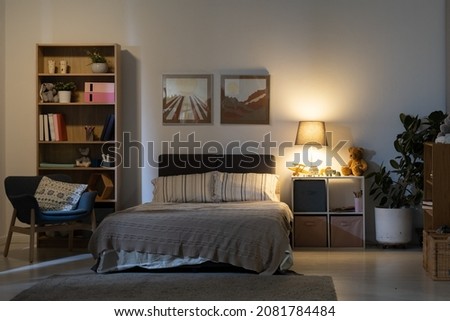 Cozy bedroom interior at night: stripped bed linen, nature pictures on wall, wooden shelves and switched lamp on bedside table
