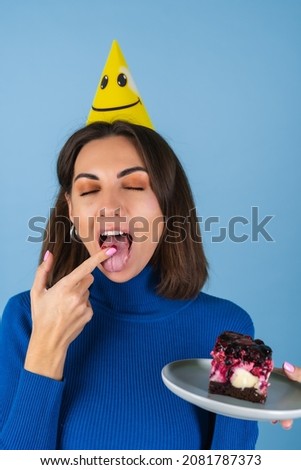 Young woman in golf on a blue background celebrates a birthday, holds a piece of cake, happy, licks her finger with pleasure