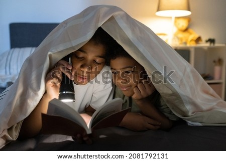 Black kids lying on bed and hiding under duvet while reading book with flashlight at night