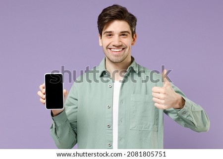 Young fun smiling man 20s in mint shirt white t-shirt using mobile cell phone blank screen workspace area show thumb up gesture isolated on purple background studio portrait. People lifestyle concept.