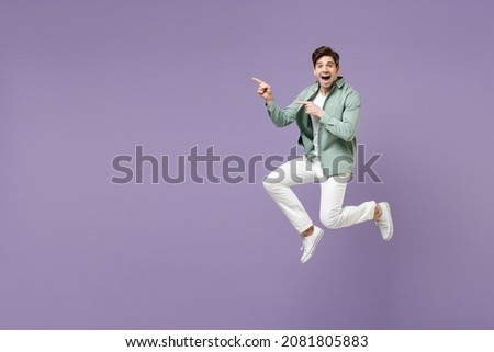 Full length fun excited cool young man in casual mint shirt white t-shirt jump high point index finger aside on copy space area mock up isolated on purple violet background. People lifestyle concept.