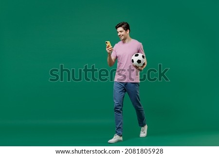 Full size body length fun young man fan in pink t-shirt cheer up football sport team hold hand soccer ball mobile cell phone watch tv live stream scream isolated on dark green color background studio