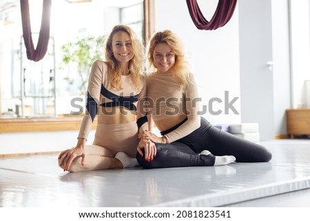 Two sporty young women smiling at camera while resting after aerial yoga workout in studio