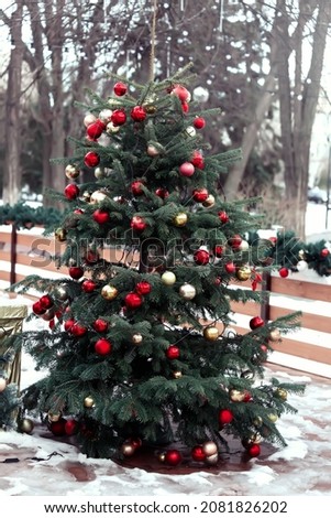 An elegant Christmas tree decorated with red and gold balloons stands in the park on the melted snow on a sunny winter day. Christmas card. New Year's street decor.
