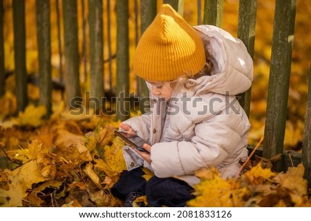 Stylish child sits on the ground in the leaves outdoors, holding a smartphone in his hands, watching cartoons. A girl uses a modern gadget for fun, playing different games online.