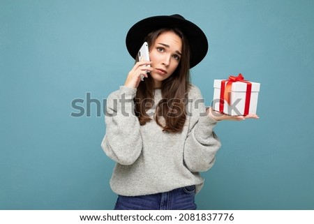 Photo of beautiful unhappy young brunette woman isolated over blue background wall with copy space for text wearing black hat and grey sweater holding white gift box with red ribbon talking on mobile