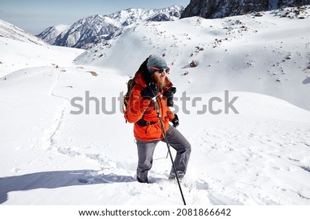 Man with backpack in red jacket climbing at the snow hill with beautiful mountains against blue sky in Almaty, Kazakhstan 