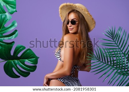 Back view of beautiful attractive young woman wears in bikini, straw hat and sunglasses who posing between tropical plants isolated on purple studio background 