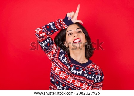 brunette caucasian girl wearing knitted sweater christmas over red background making fun of people with fingers on forehead doing loser gesture mocking and insulting.