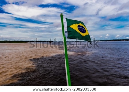 Flag of Brazil hoisted on mast. And the meeting of the Negro River with the Solimões forming the Amazon River. Royalty-Free Stock Photo #2082033538