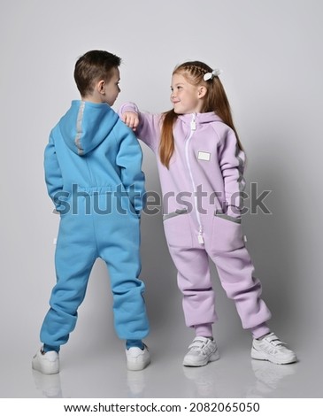 Two cute kids in blue and pink jumpsuits are standing next to and looking at each other, boy with his back to us and girl with her face over light background